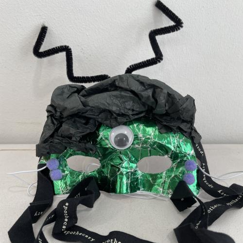 mask decorated with green shiny paper, black tissue paper and a single googly eye