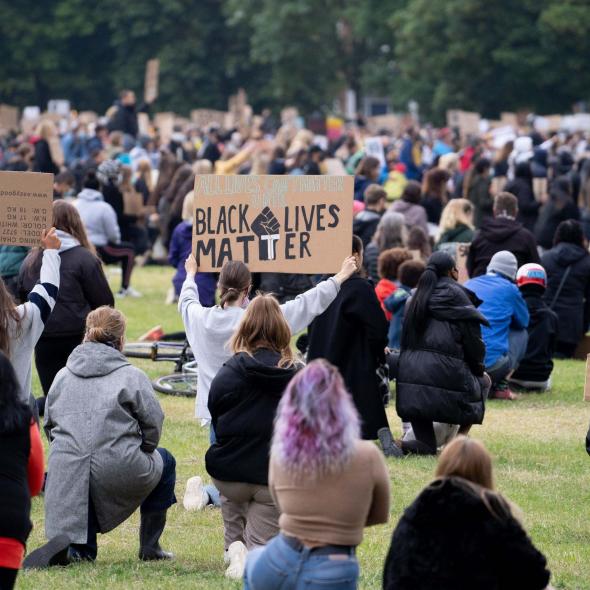 A crowd of people kneeling in a field shown from behind. One person is holding up a sign which says Black Lives Matter