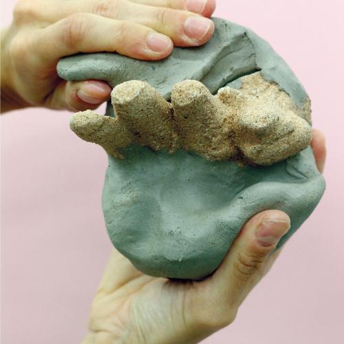 Two hands holding a sculpture made of green clay with stones sticking out