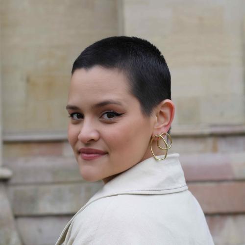 Photograph of a woman with short cropped black hair and a gold earring looking over her shoulder at the camera