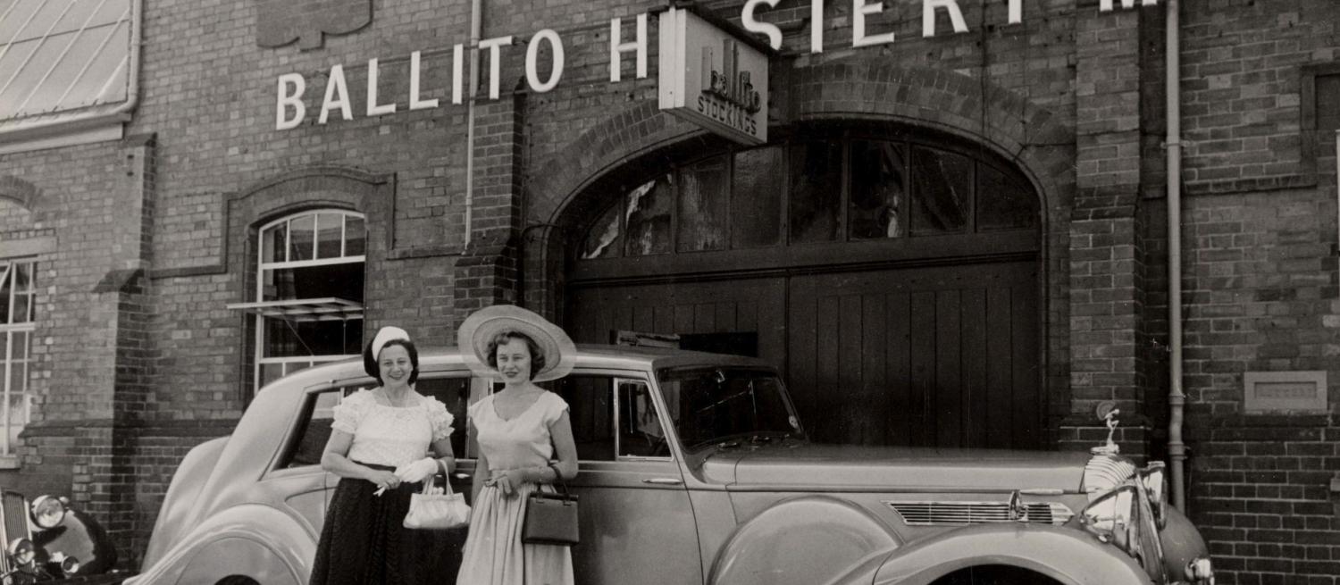 A black and white photograph showing two ladies in 1950s dresses and large hats standing in front of a car by the door of a building. Over the door it says Ballito Hosiery Mill
