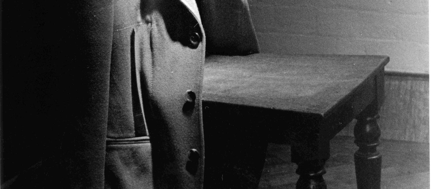 Black and white photograph of a jacket hanging on the back of a wooden chair. The left hand edge of the picture fades into a shadow.