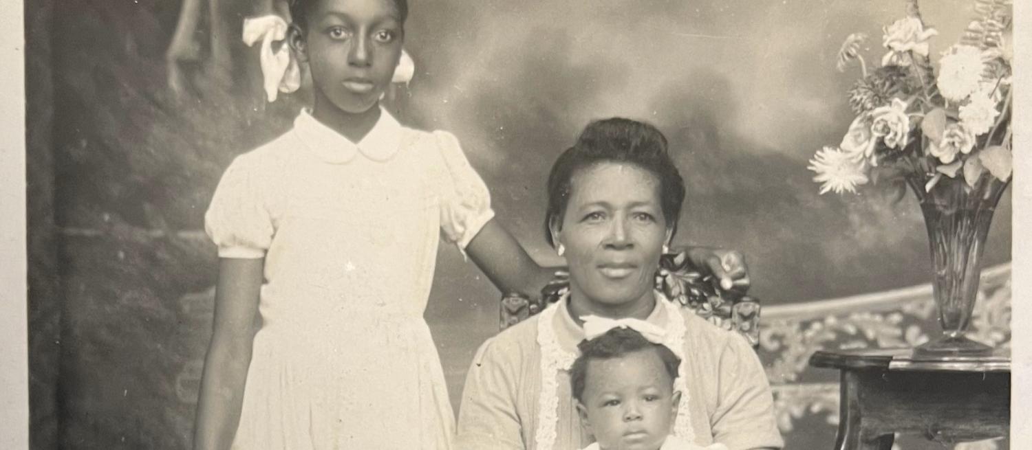 A black and white group portrait showing three Black sitters in an interior. A baby sits on a woman's knee and a child stands to one side. 