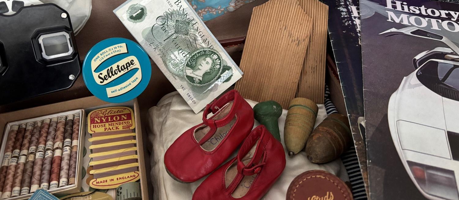 Colour photograph of a selection of vintage and nostalgic objects including wooden spinning tops, old magazines, old fashioned textiles, tint red baby shoes, a viewfinder toy and more.