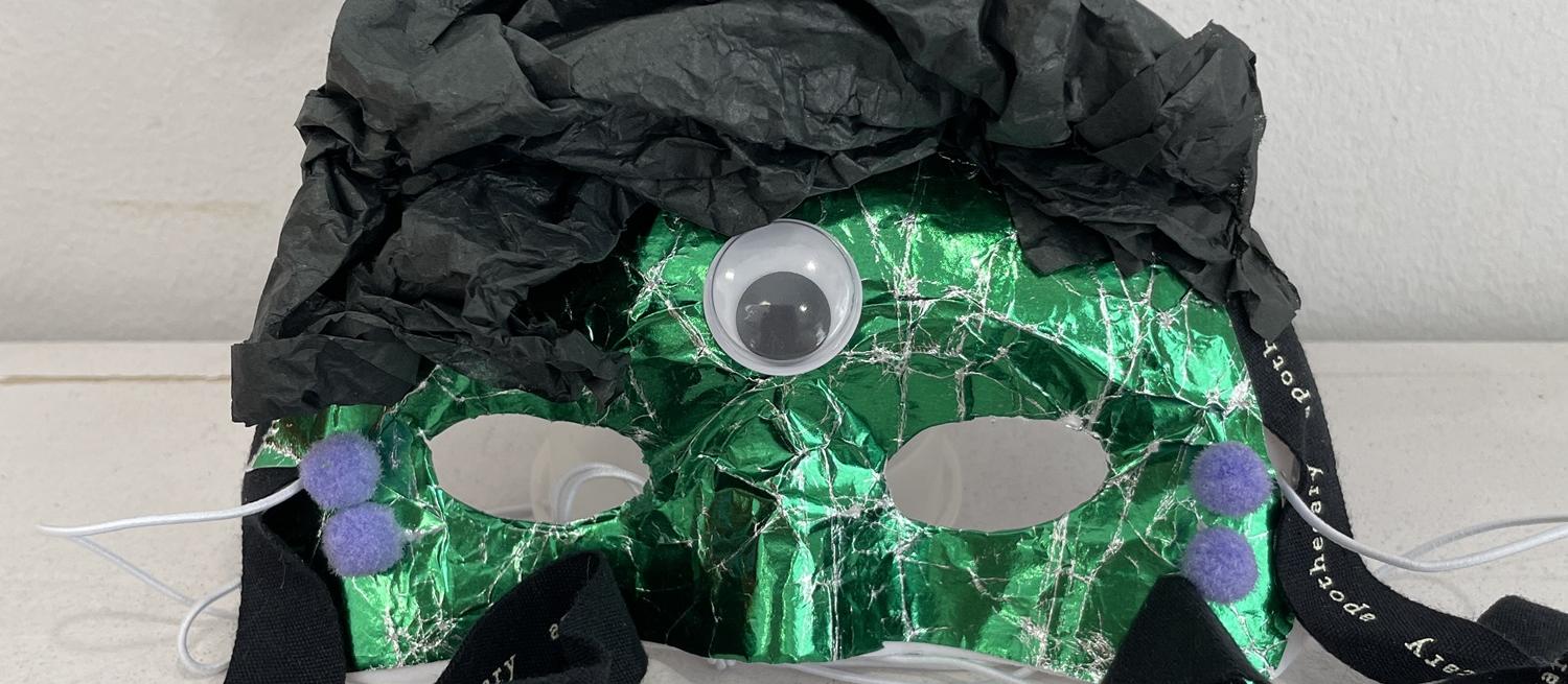 mask decorated with shiny green paper, black tissue paper and a single googly eye