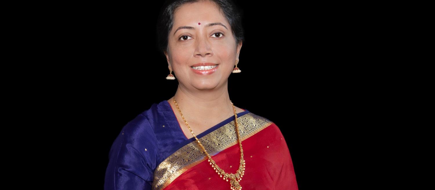A posed portrait of a lady with Indian heritage wearing a saree and gold necklace