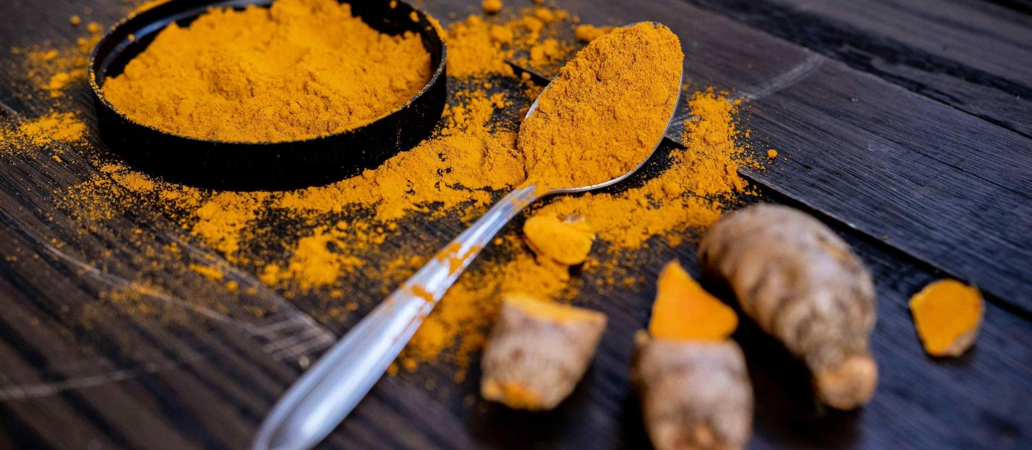 Ground turmeric overflows from a black shallow dish and a silver tea spoon. Three larger chunks of chopped turmeric sit to the right hand side. 