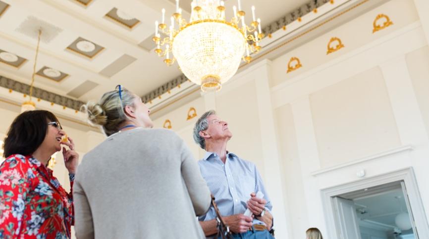 Three museum visitors, two women and one man, look up at a large, sparkling crystal chandelier hanging from a high, white ceiling with gold decorative trim.