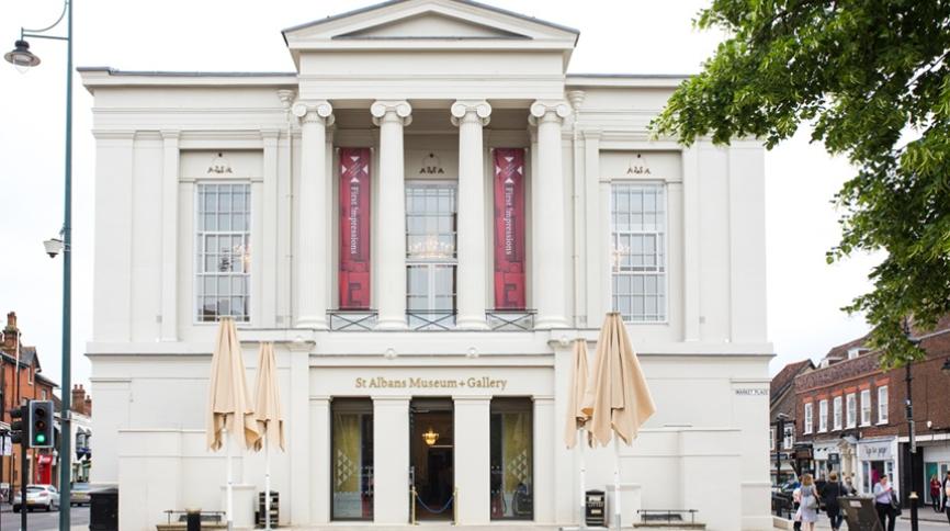 A white classical building with four columns and a pediment. Banners reading "first impressions" hang between the columns, and the sign "St Albans Museum + Gallery" is above the entrance. Two folded beige umbrellas stand on the paved area in front.