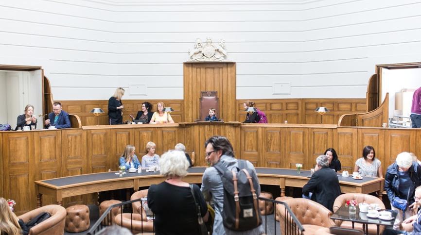 A former courtroom, now a cafe, with people seated at tables and in brown leather armchairs. The room features curved wooden paneling, a raised judge's bench with a crest, and a high white wall.