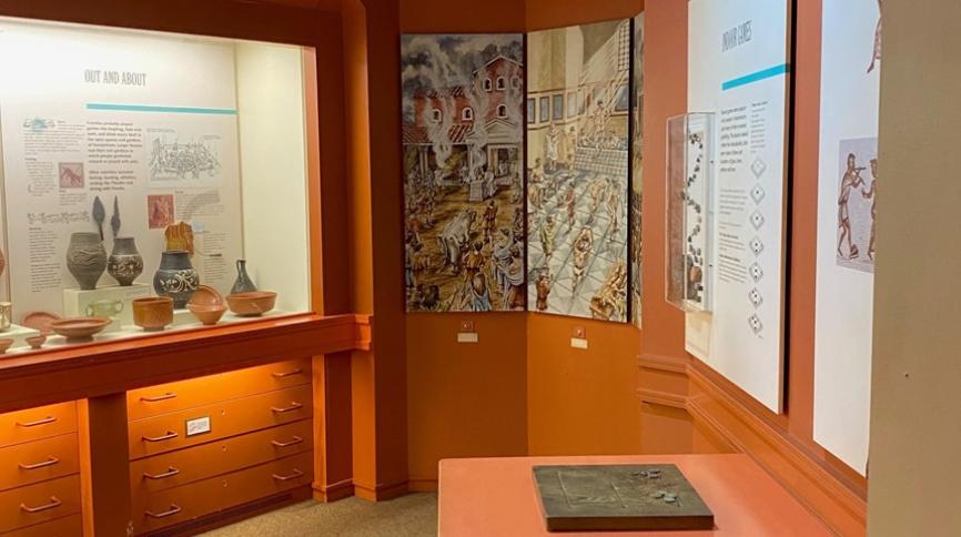 A room with a display case and picture of a Roman bathhouse on the wall. In the foreground, there is a wooden boardgame on a table.
