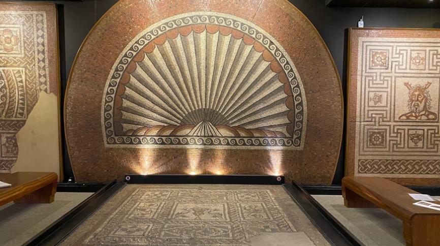 A large semi-circular mosaic with a fan-shaped sea shell, surrounded by a border of black and white waves. In front on the ground is another mosaic, in the centre is two dophins and a cup surrounded by a chain like pattern. On the right hand side we can see half of a mosaic featuring a horned-god in the centre.