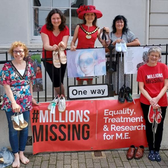 A colour photograph of members of the Herts ME Group standing in front of the railings at the St Albans Museum and Gallery with printed artworks and a banner for the 'Missing Millions' Campaign. 