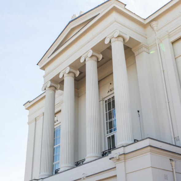 A light-colored classical building features six tall, fluted columns supporting a triangular pediment. Large windows are visible on the facade, under a clear, light blue sky.