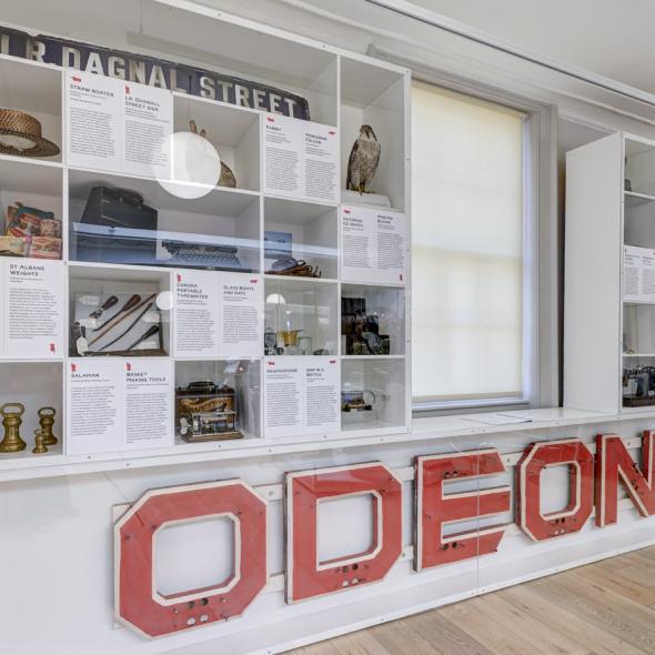 Museum display case containing large ODEON cinema sign, Victorian straw boater hat, peregrine falcon taxidermy and other museum collection ephemera