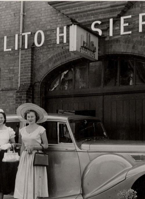 A black and white photograph showing two ladies in 1950s dresses and large hats standing in front of a car by the door of a building. Over the door it says Ballito Hosiery Mill