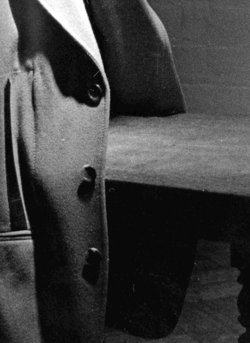 Black and white photograph of a jacket hanging on the back of a wooden chair. The left hand edge of the picture fades into a shadow.