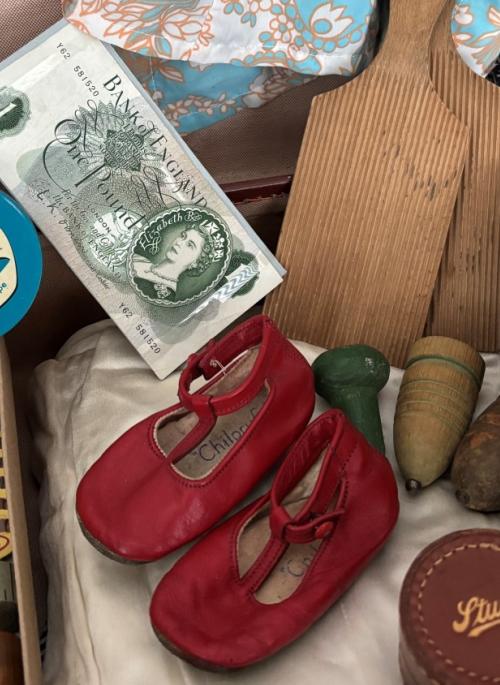 Colour photograph of a selection of vintage and nostalgic objects including wooden spinning tops, old magazines, old fashioned textiles, tint red baby shoes, a viewfinder toy and more.