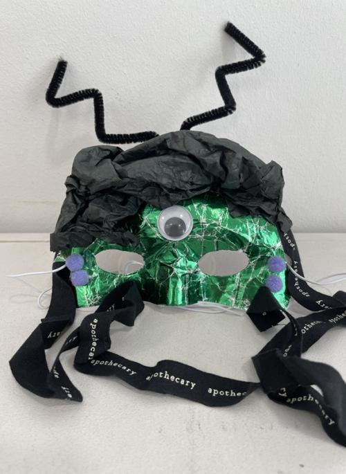 mask decorated with shiny green paper, black tissue paper and a single googly eye